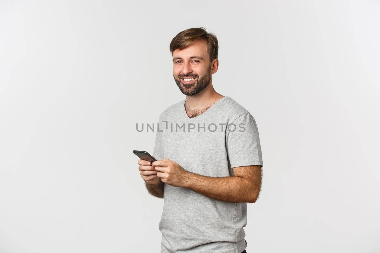 Portrait of handsome bearded guy with a smartphone, smiling pleased at camera, standing over white background by Benzoix