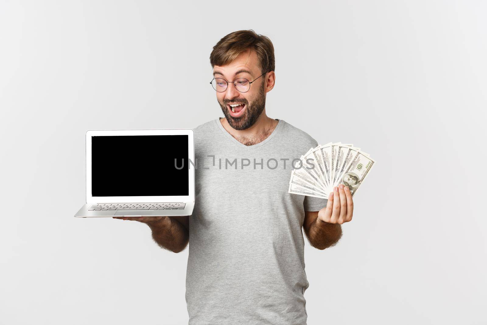Handsome excited male freelancer showing laptop screen and money, standing over white background.