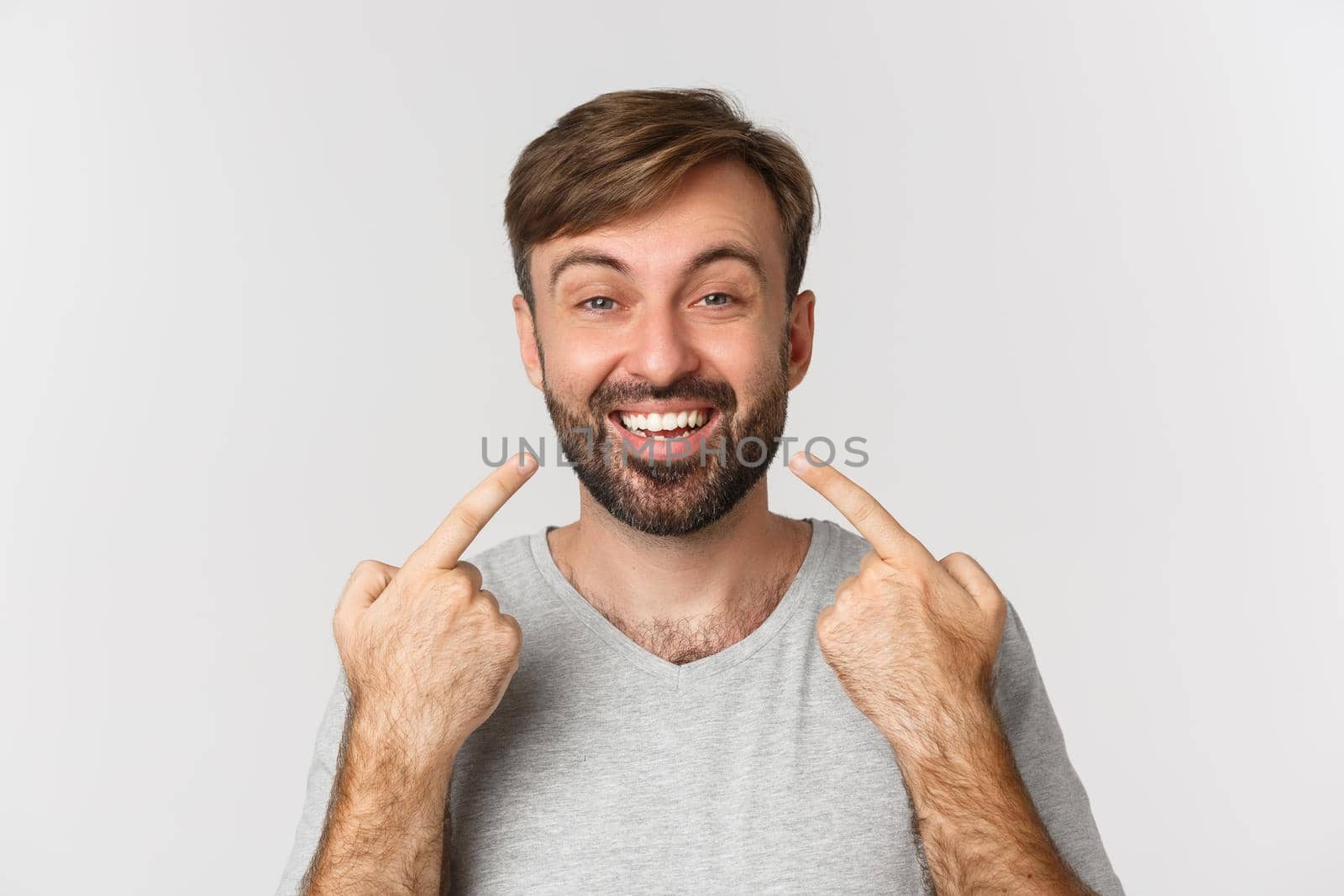 Close-up of handsome smiling man recommending dental clinic, pointing at his perfect white teeth, standing over white background.