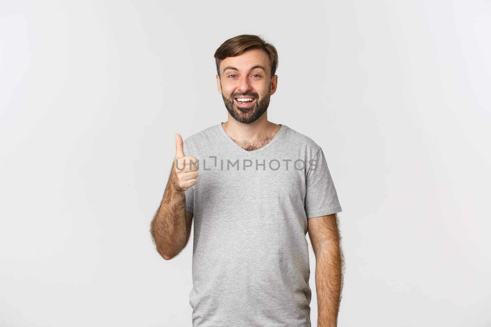 Image of satisfied smiling man in gray t-shirt, showing thumbs-up in approval, praising good choice, white background.