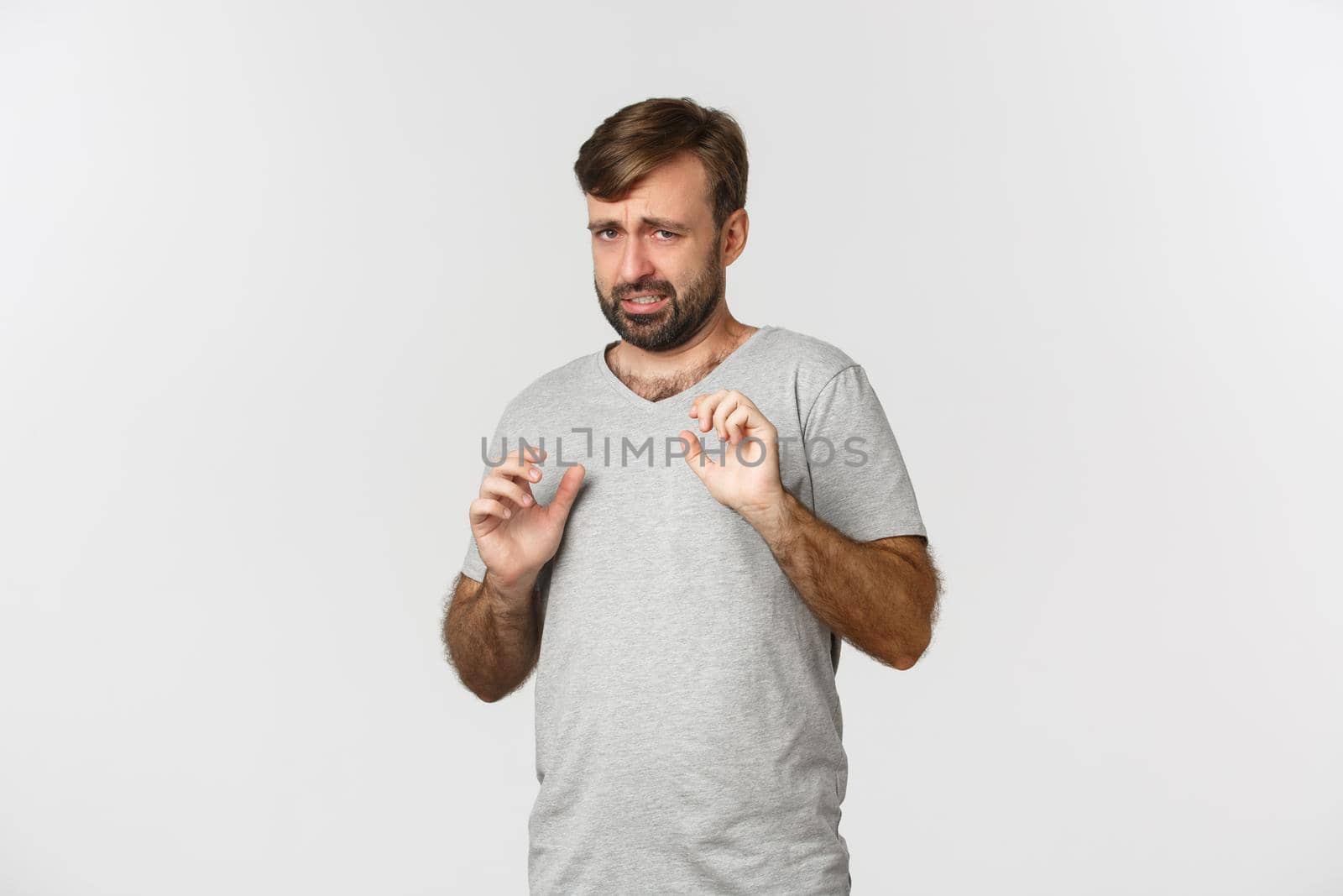 Portrait of timid and scared man in gray t-shirt, raising hands to defend himself, looking frightened, standing over white background.