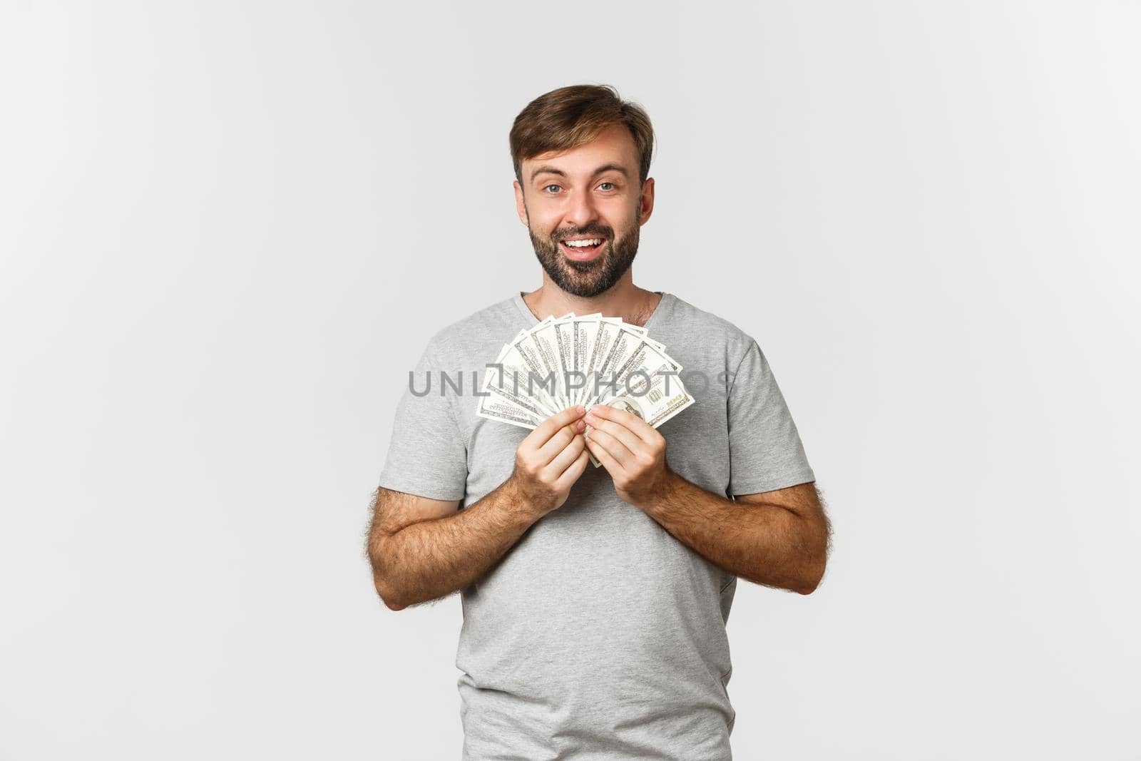 Smiling man with beard, wearing gray t-shirt, showing money, winning prize, standing over white background.