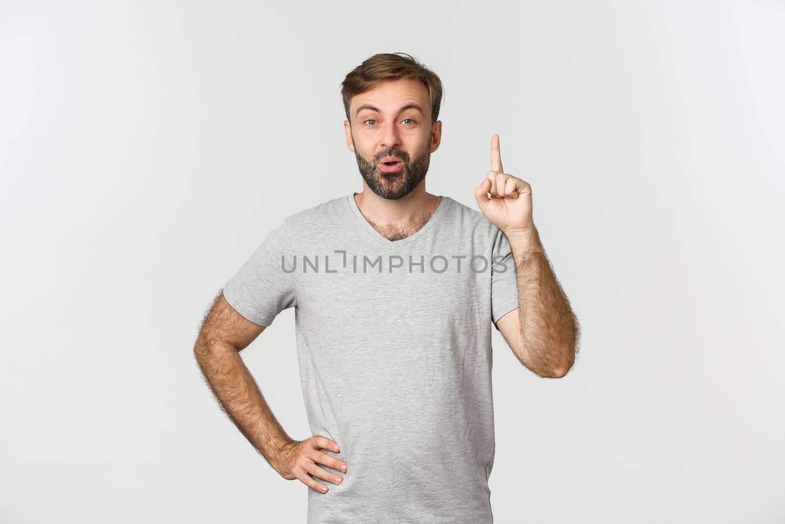 Portrait of handsome bearded man in gray t-shirt, raising index finger, having suggestion, have an idea, standing over white background.