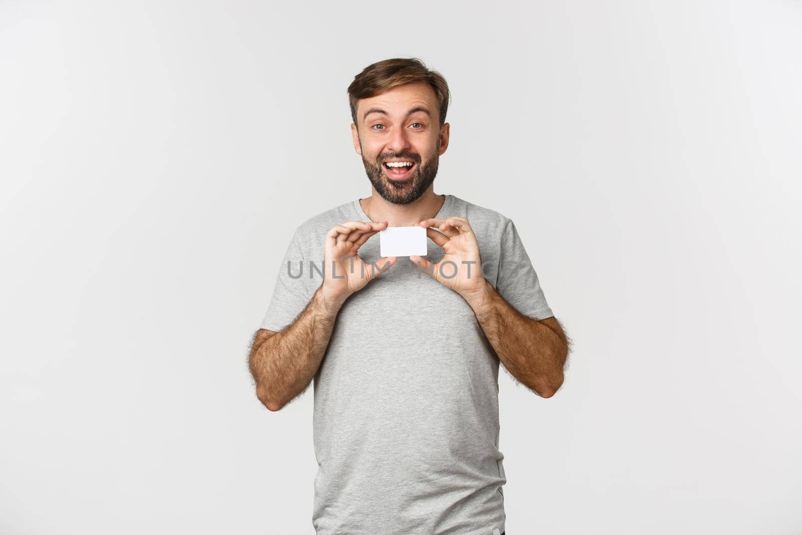 Cheerful caucasian man in gray t-shirt, showing credit card and smiling, excited for shopping, standing over white background.