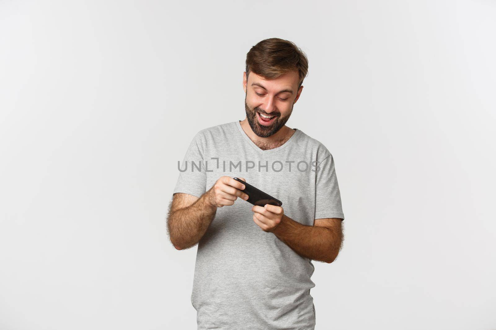 Image of adult bearded man in gray t-shirt, playing mobile games and having fun, standing over white background by Benzoix