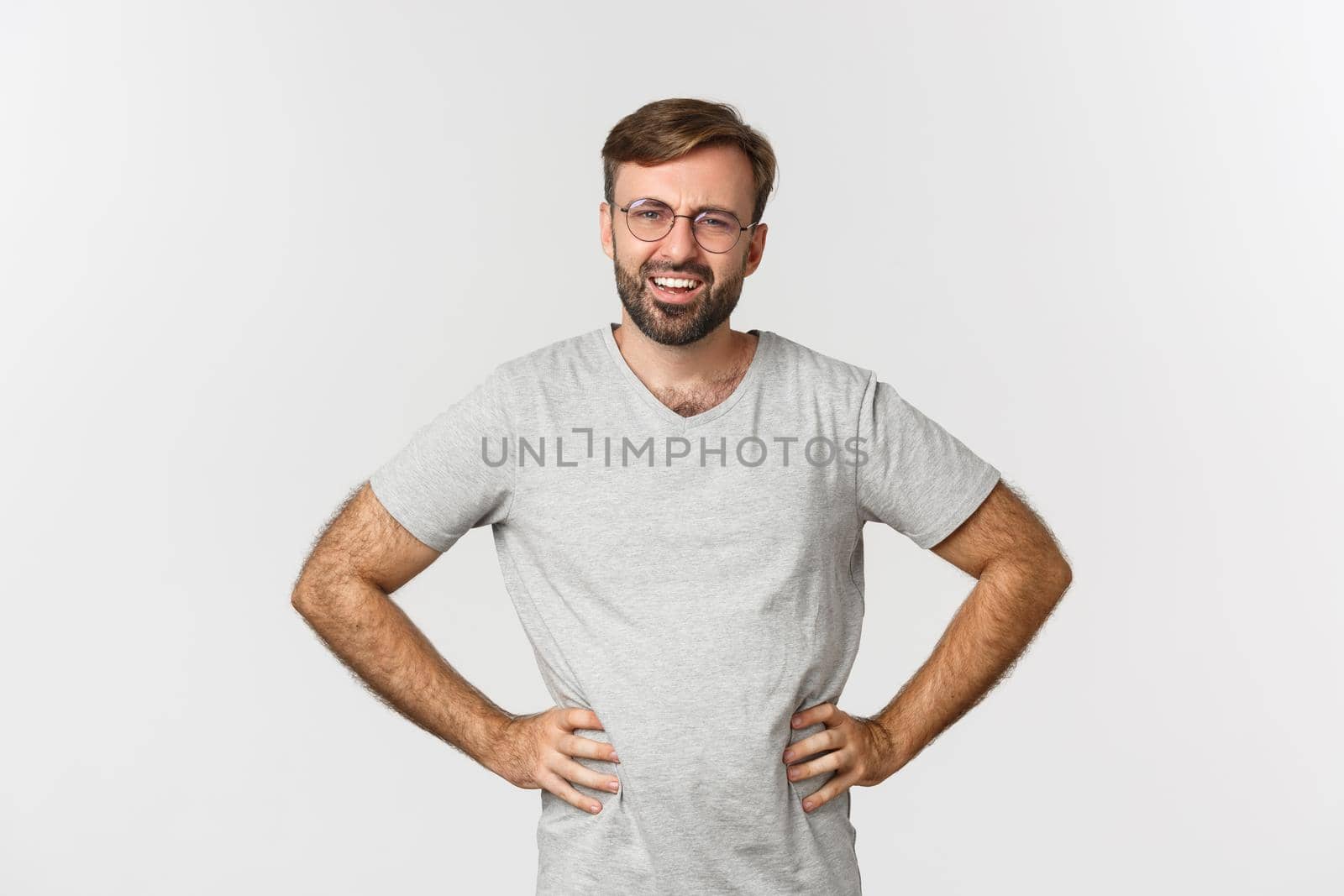 Image of confused bearded man in glasses and gray t-shirt, looking at something strange, frowning perplexed, standing over white background.