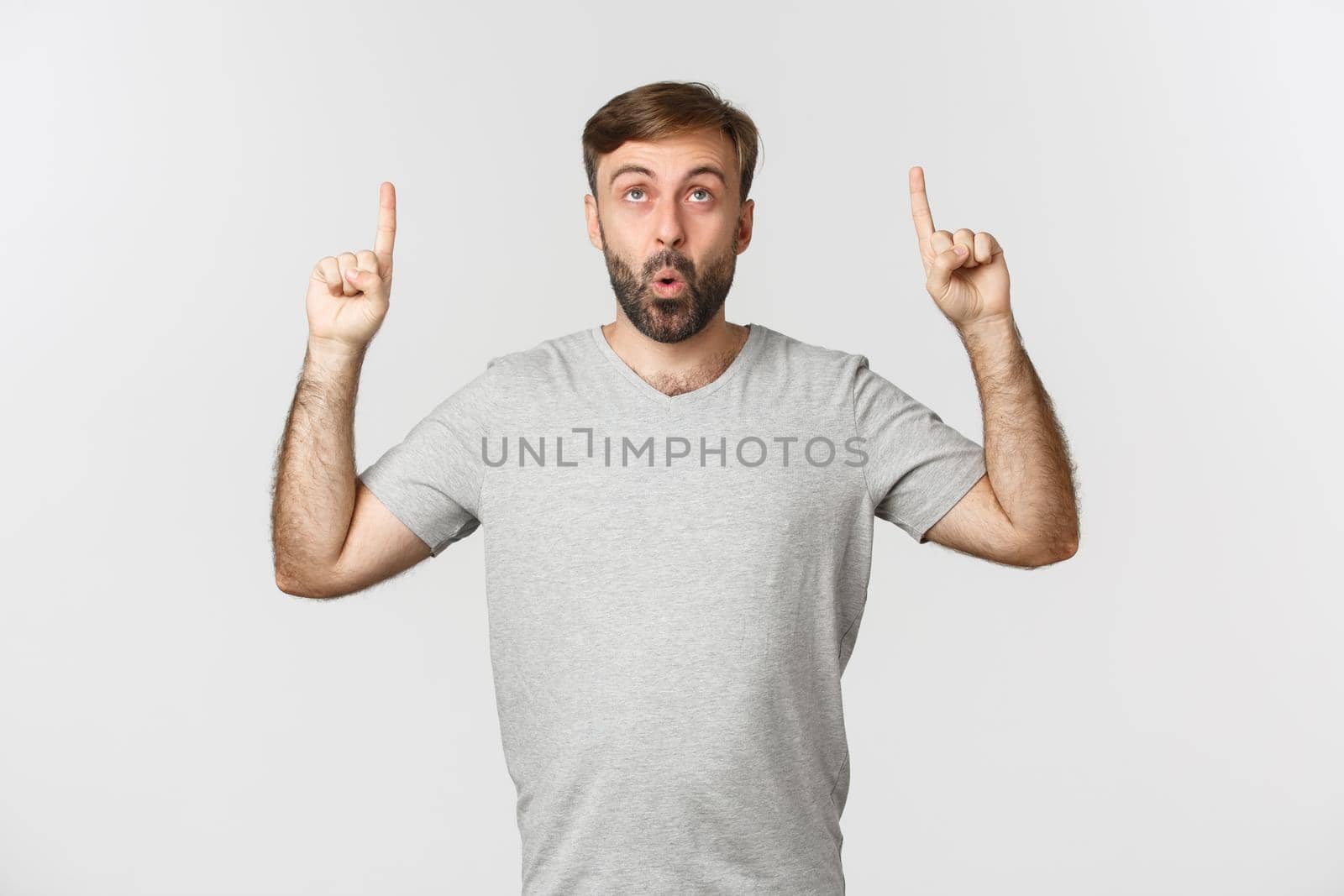 Portrait of adult caucasian guy with beard, wearing gray t-shirt, looking impressed at something cool, pointing fingers up, showing promo, standing over white background by Benzoix