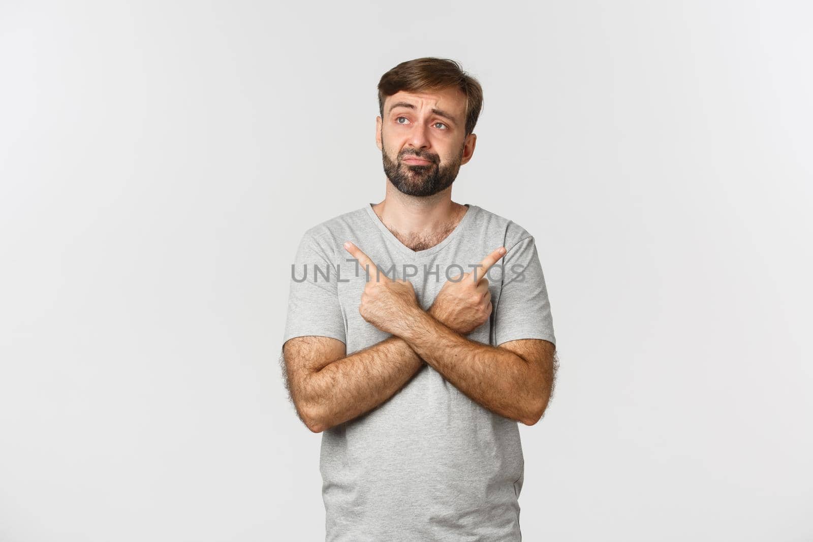 Portrait of indecisive beaded man in gray t-shirt, making decision, pointing fingers sideways at two choices, standing over white background.