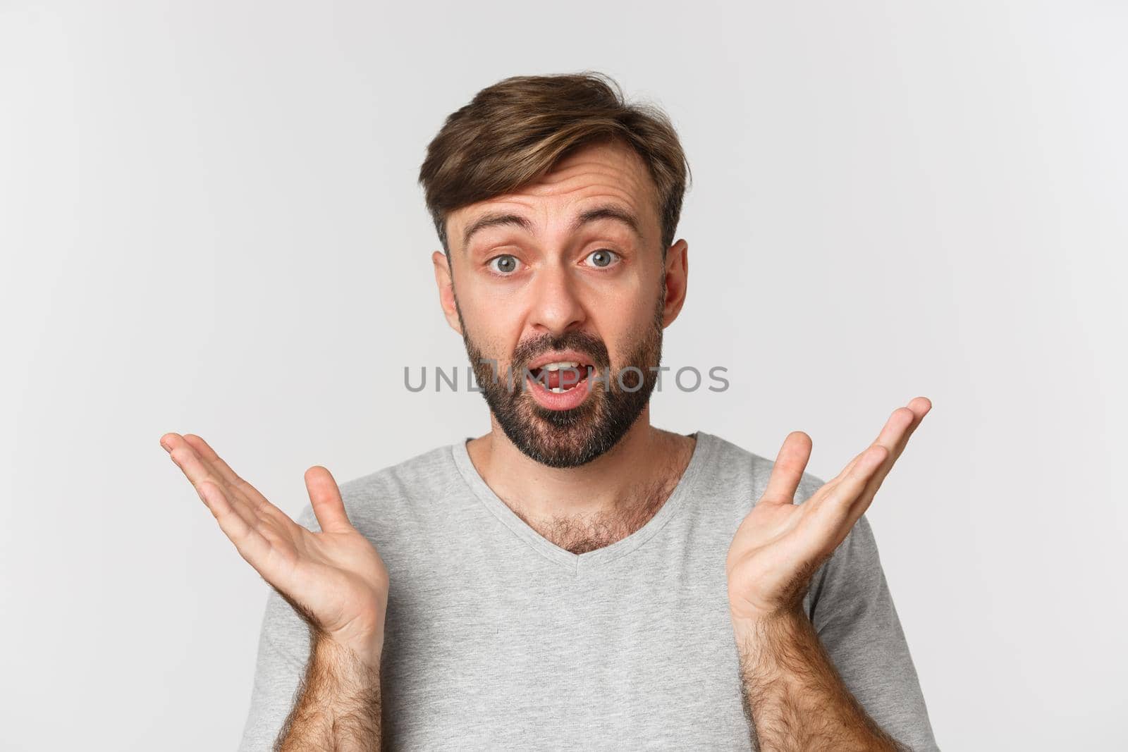 Close-up of surprised handsome man with beard, spread hands sideways and look in awe, standing over white background.