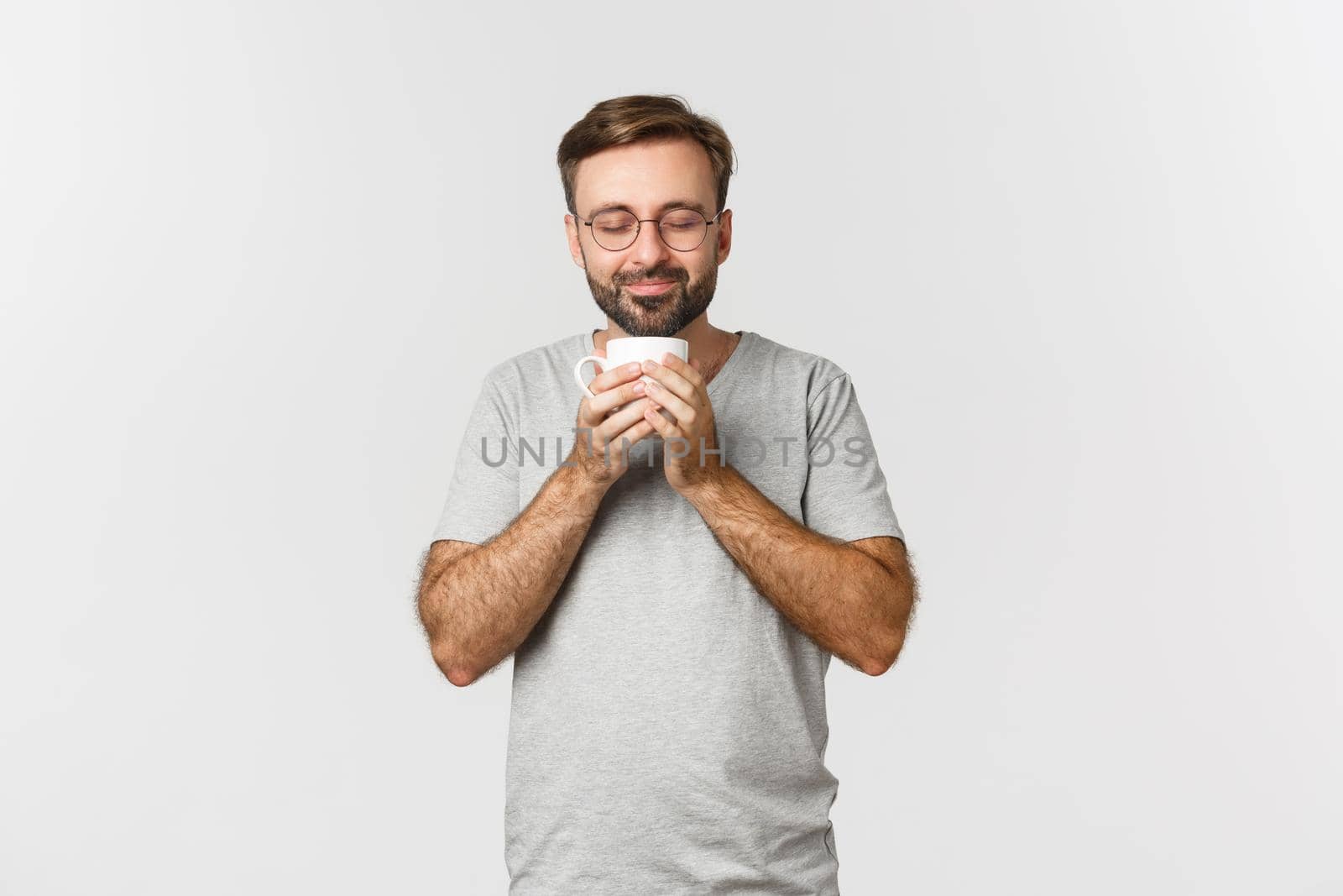 Portrait of happy smiling man in glasses and casual t-shirt, close eyes and smelling coffee in mug, standing over white background.