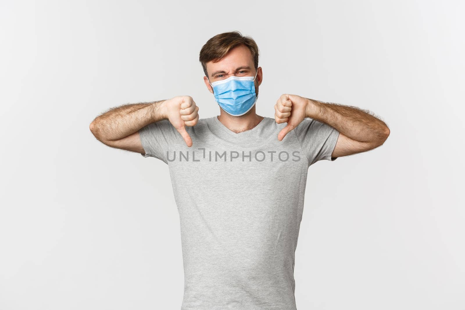 Concept of pandemic, covid-19 and social-distancing. Portrait of man hate wearing medical mask, complaining and showing thumbs-down, standing over white background.