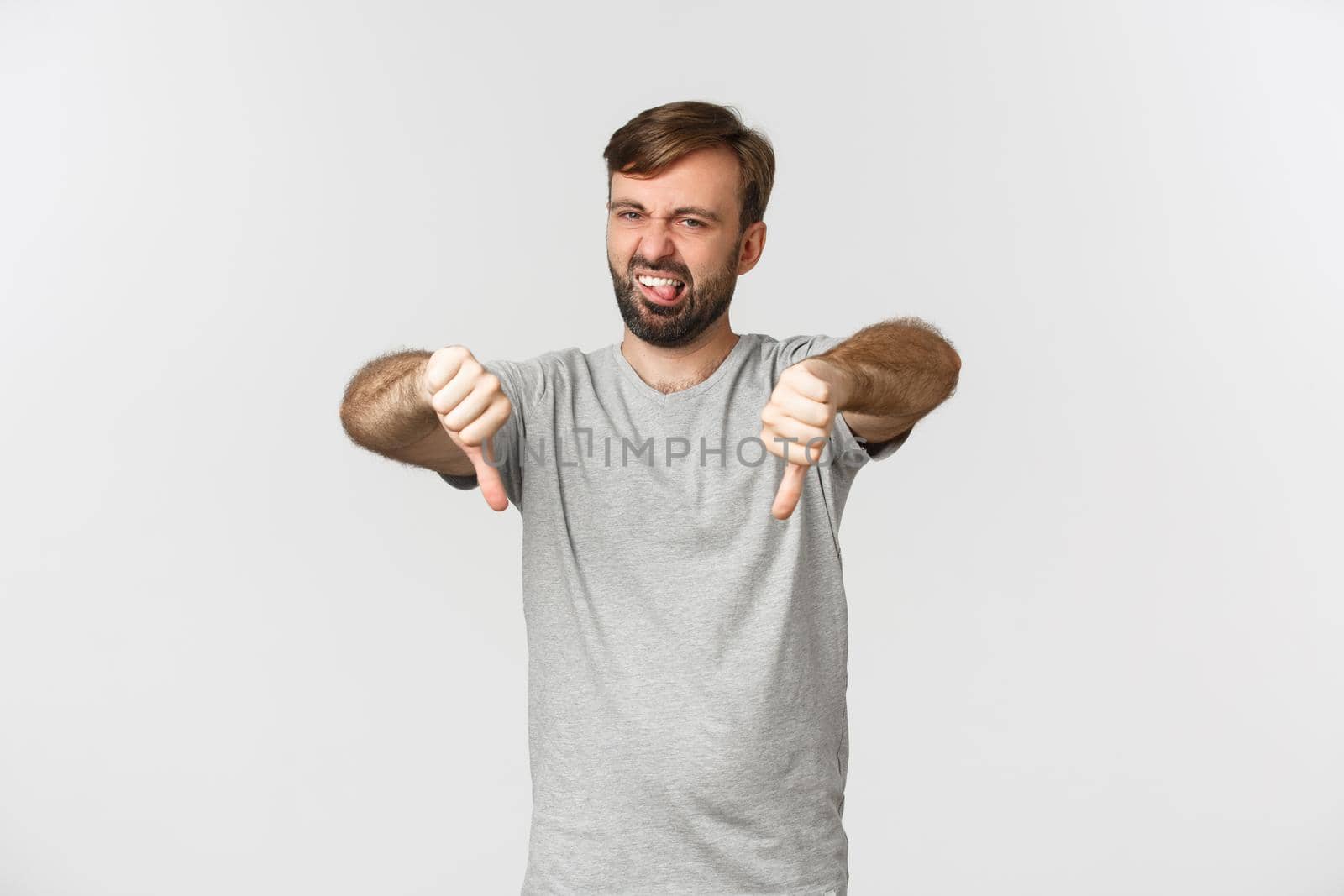 Portrait of skeptical bearded man in casual t-shirt, showing thumbs-down and looking disappointed, disapprove something bad, standing over white background.