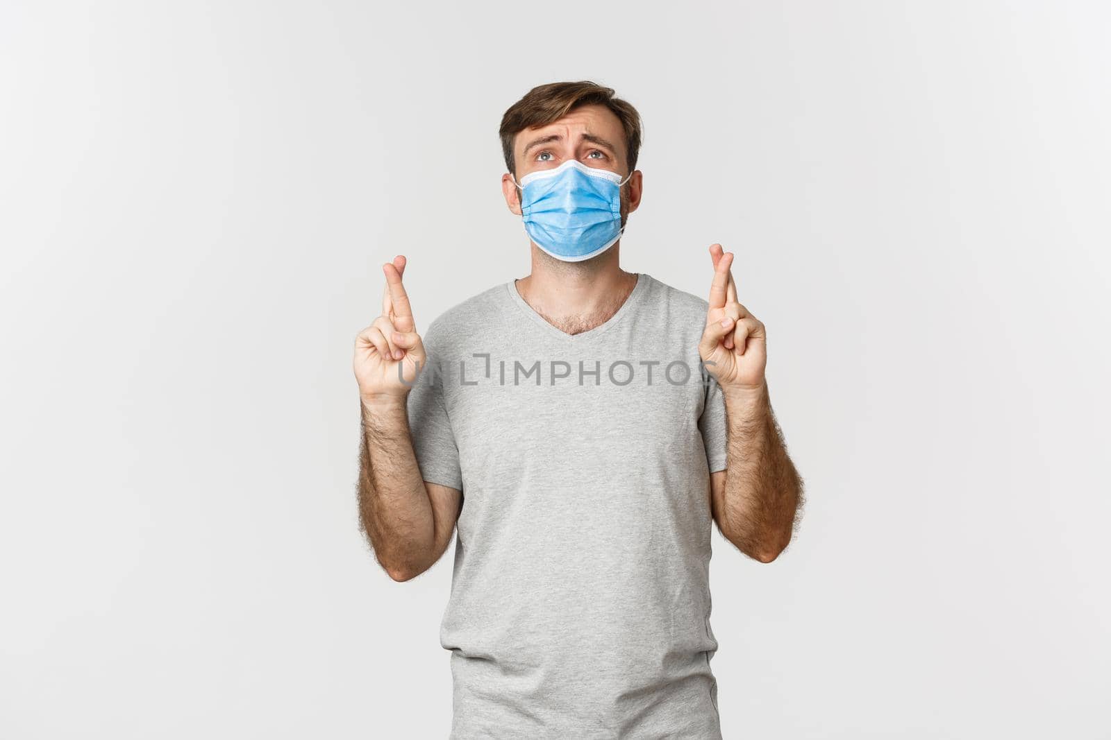 Concept of pandemic, covid-19 and social-distancing. Hopeful worried guy in medical mask, cross fingers for good luck and begging god, looking up desperate, standing over white background.