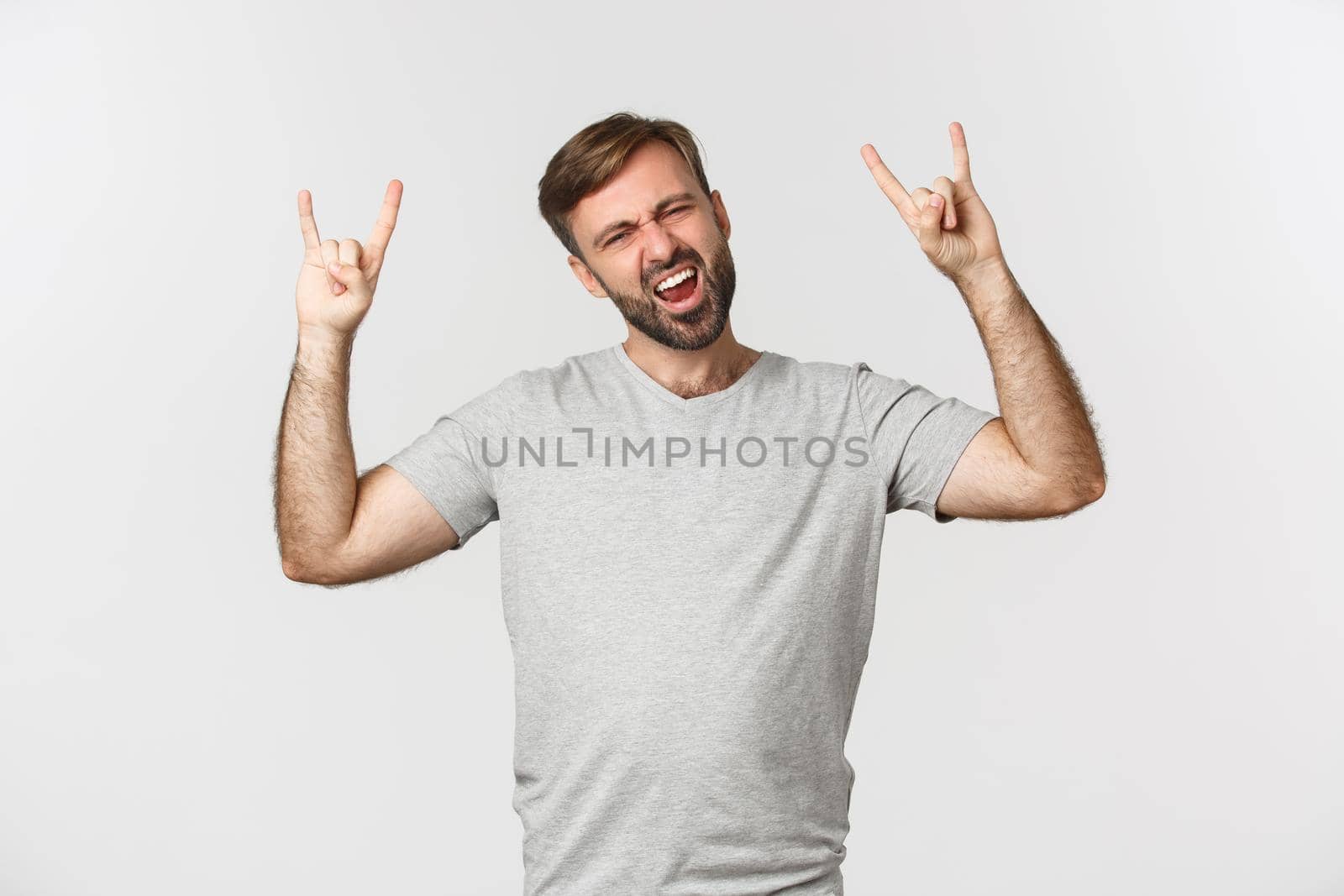 Portrait of carefree bearded man having fun, showing rock-n-roll gesture and shouting for joy, standing over white background.
