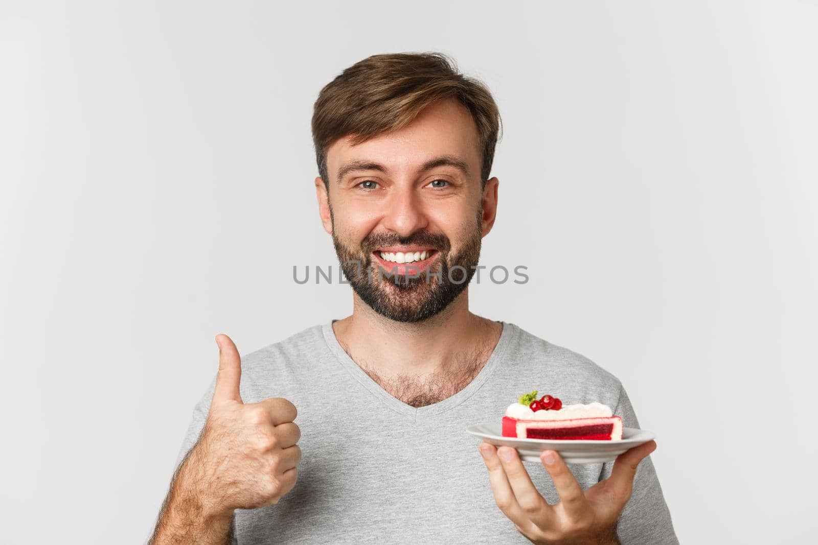 Close-up of cheerful caucasian guy with beard, wearing grey t-shirt, holding cake and showing thumbs-up, like dessert, standing over white background.