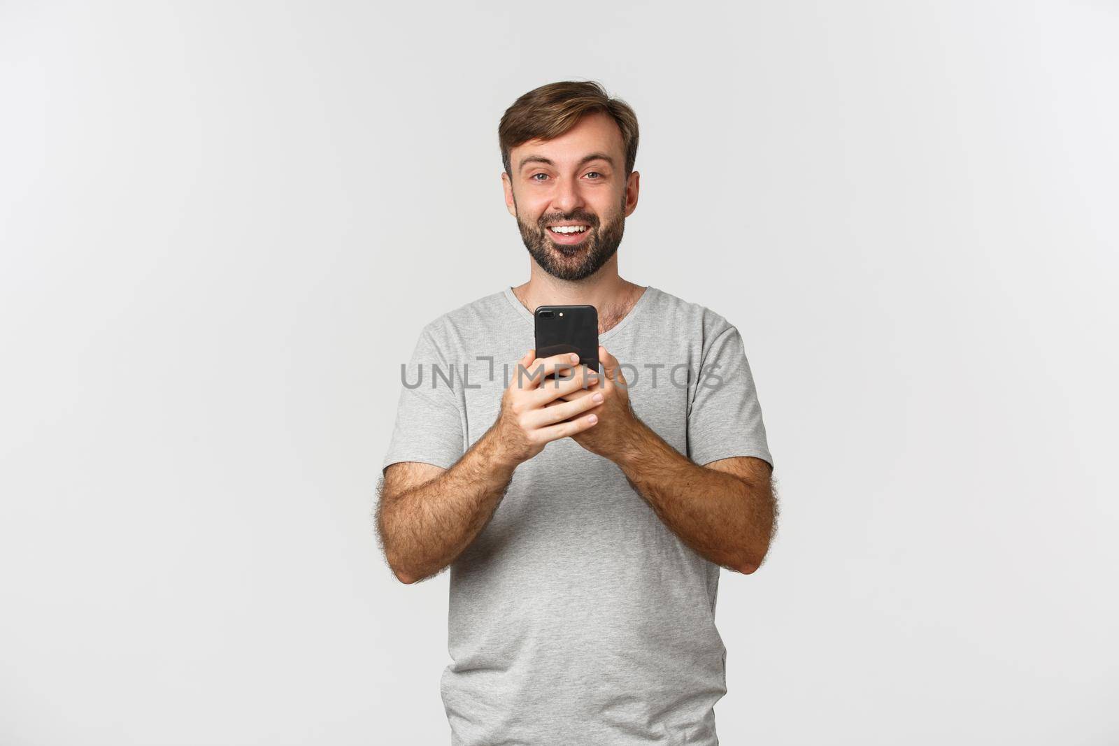 Smiling guy with beard, taking pictures or recording video on mobile phone, standing over white background.