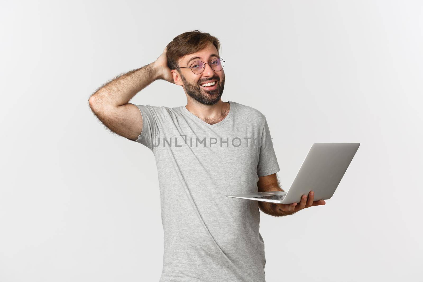 Portrait of humble handsome guy in glasses and t-shirt, working with laptop and laughing, standing over white background.