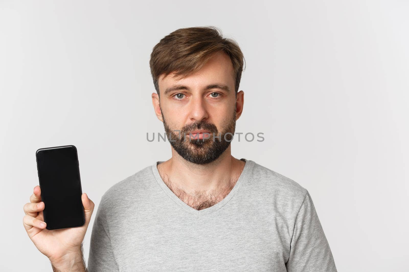 Close-up of tired caucasian guy in gray t-shirt, looking exhausted and showing smartphone screen, standing over white background.