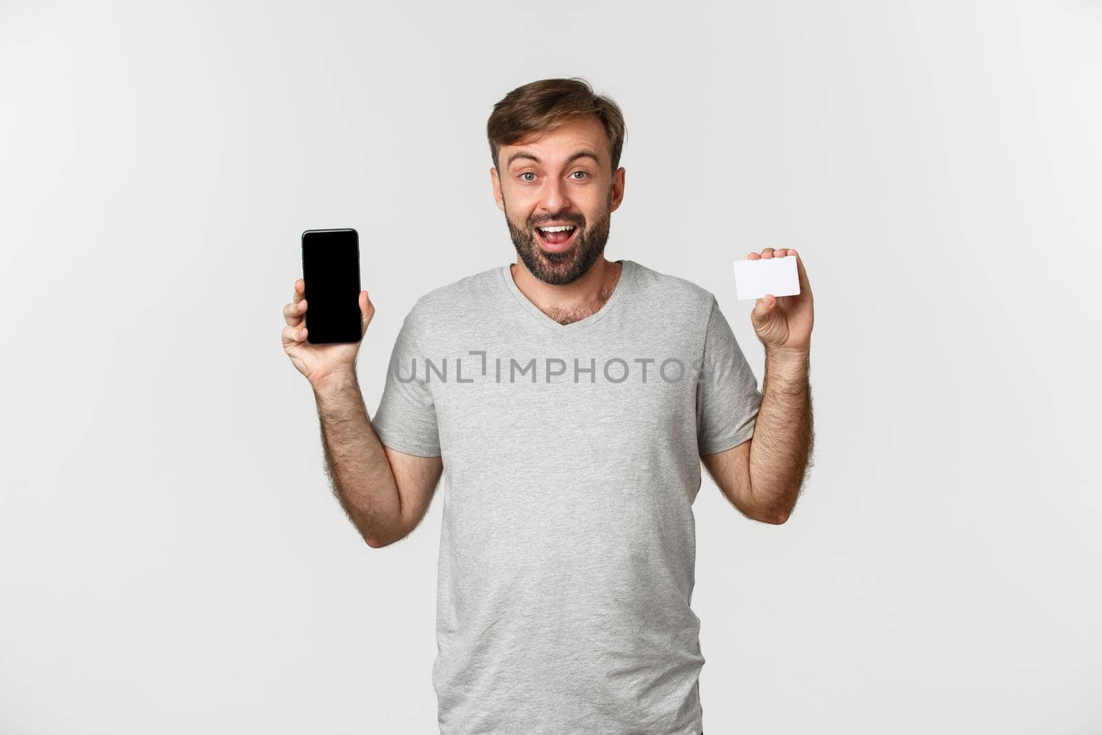 Handsome young man shopping online, holding credit card and mobile phone, showing smartphone screen, standing over white background.