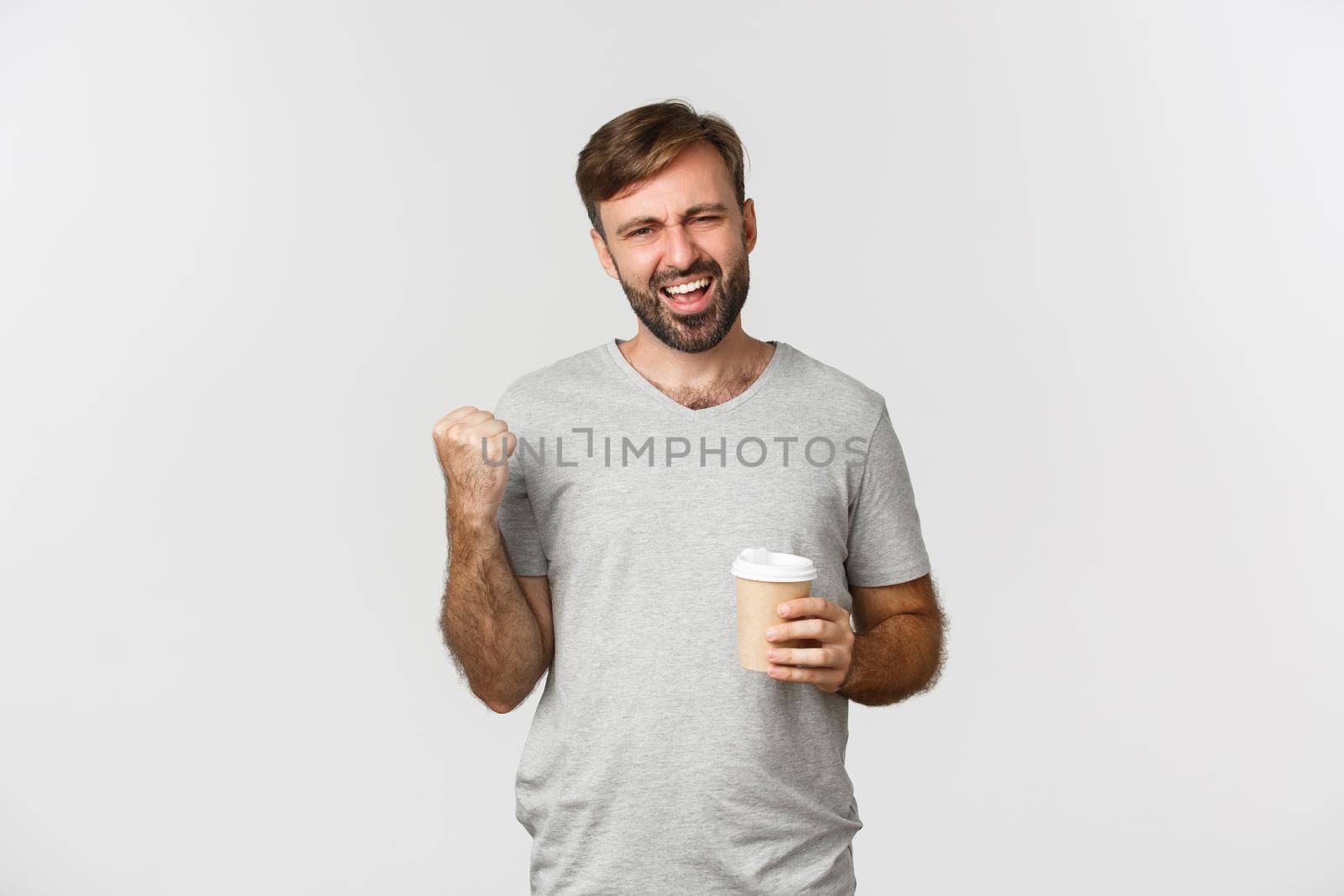 Portrait of handsome man with beard, wearing gray basic t-shirt, feeling energized after drinking coffee, making fist pump and saying yes with excitement, standing over white background.