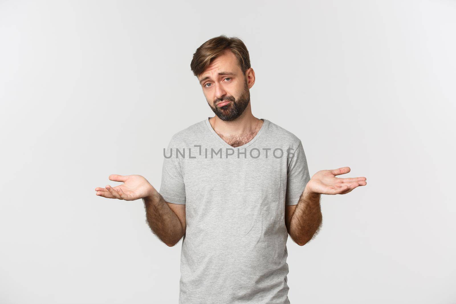 Portrait of indecisive sad guy with beard, shrugging with hands spread sideways, standing clueless over white background.