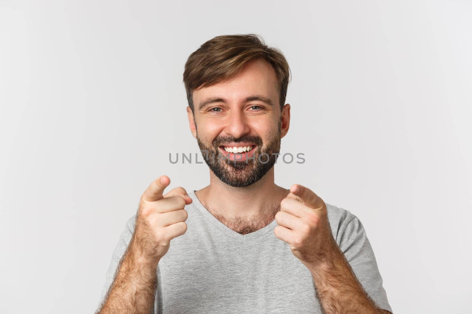 Close-up of handsome adult man with beard, smiling and pointing fingers at camera, standing in gray t-shirt over white background.