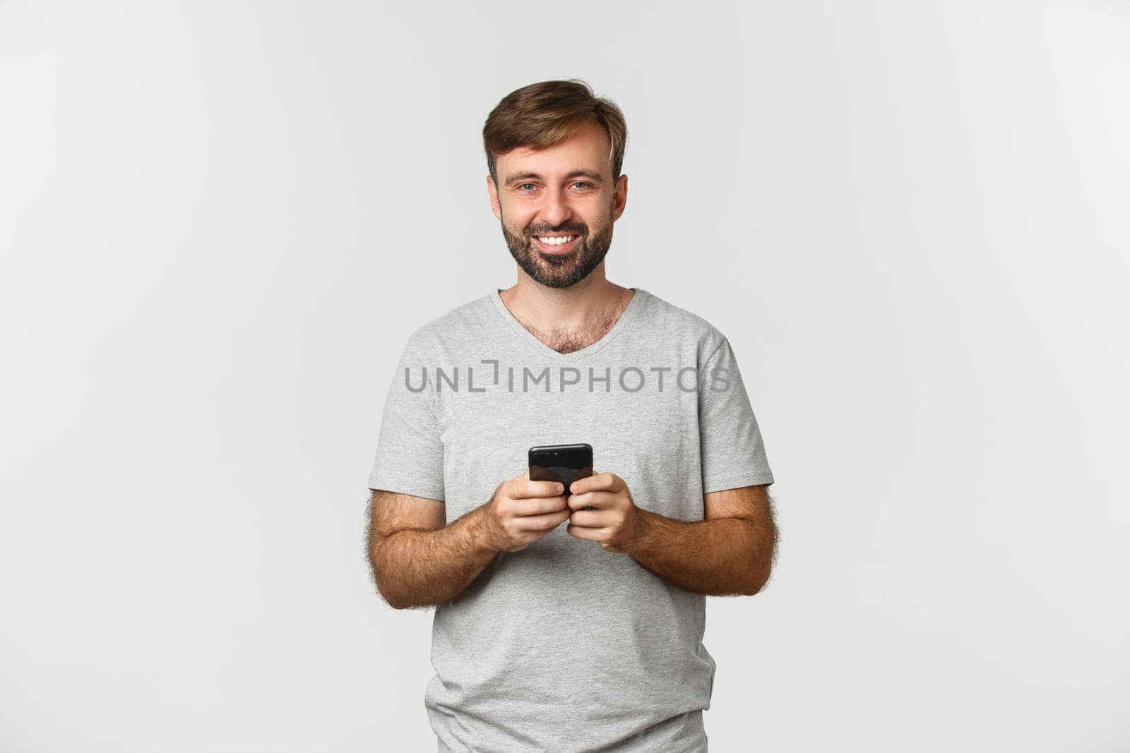Portrait of happy smiling man with beard, wearing t-shirt, using mobile phone, standing over white background.