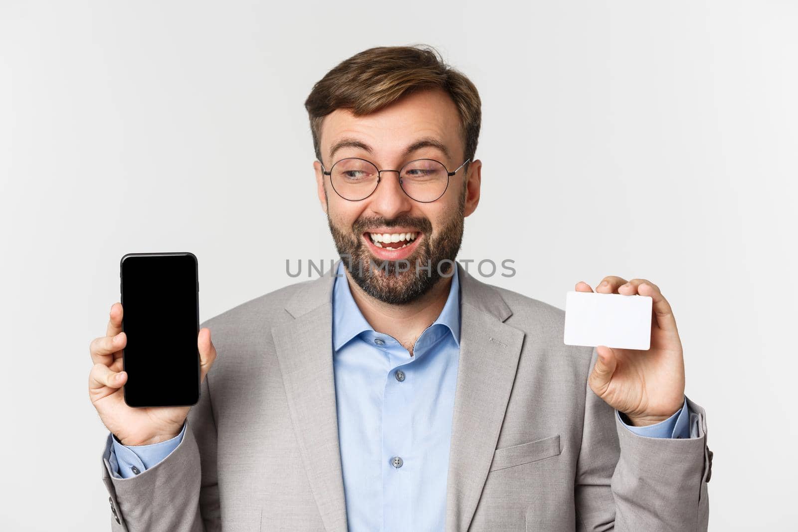 Close-up of handsome smiling male entrepreneur, wearing glasses and gray suit, showing credit card and looking excited at smartphone screen, standing over white background.