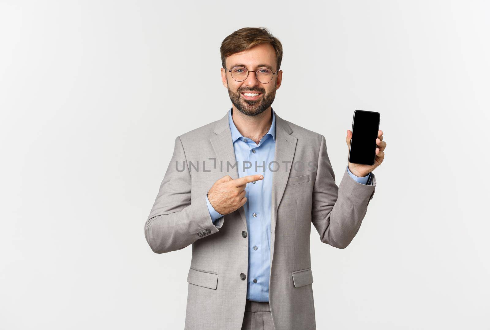 Handsome smiling businessman with beard, wearing grey suit and glasses, pointing finger at mobile phone screen, showing app, standing over white background by Benzoix