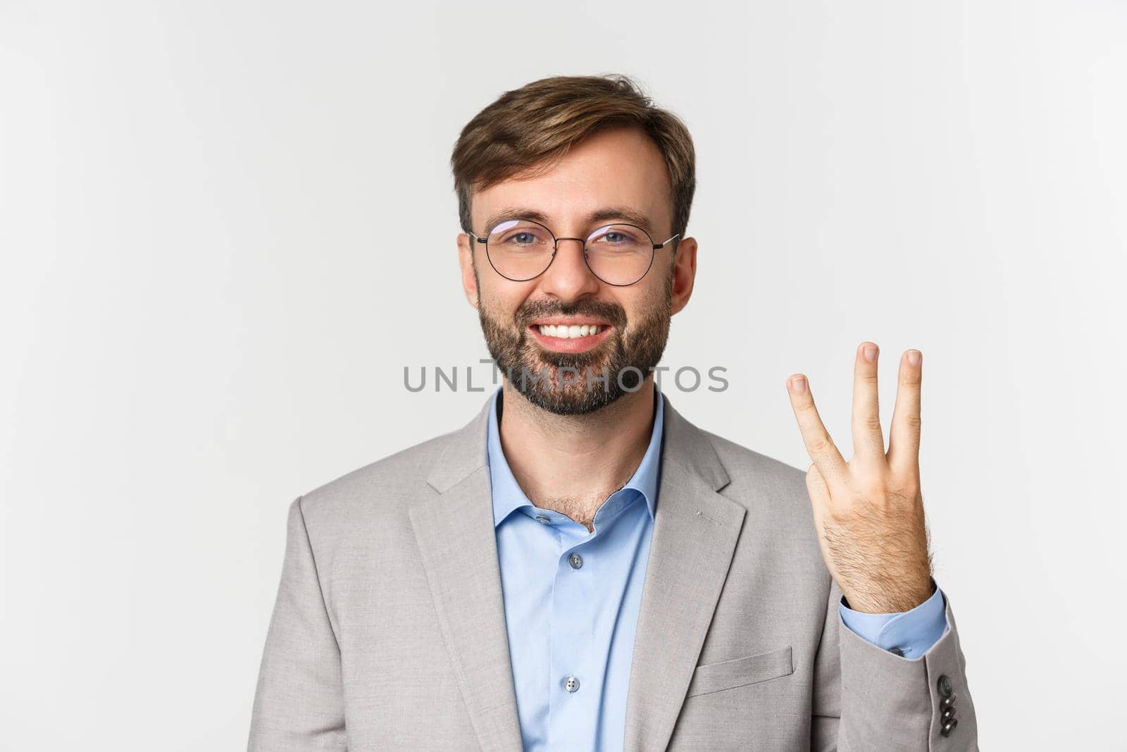 Close-up of handsome bearded man in glasses and gray suit, showing number three and smiling, standing over white background.