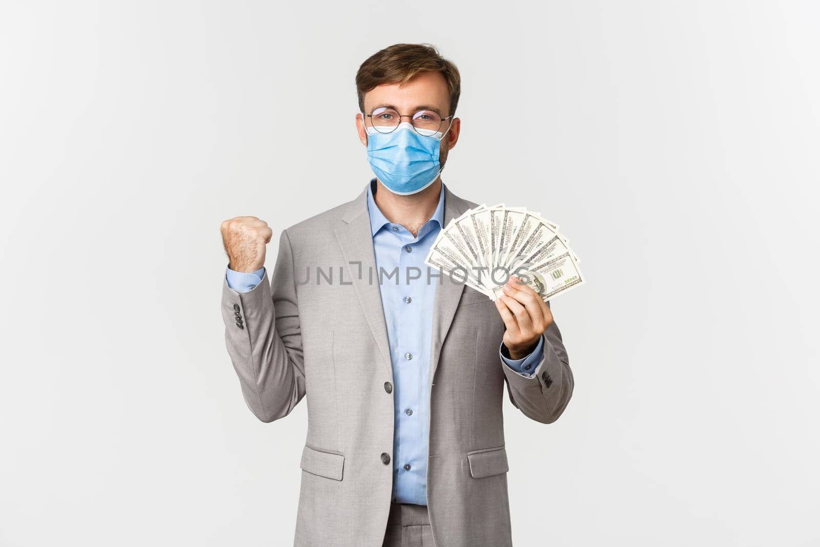 Concept of business, covid-19 and social distancing. Portrait of confident businessman in gray suit and medical mask, rejoicing and showing money, standing over white background.