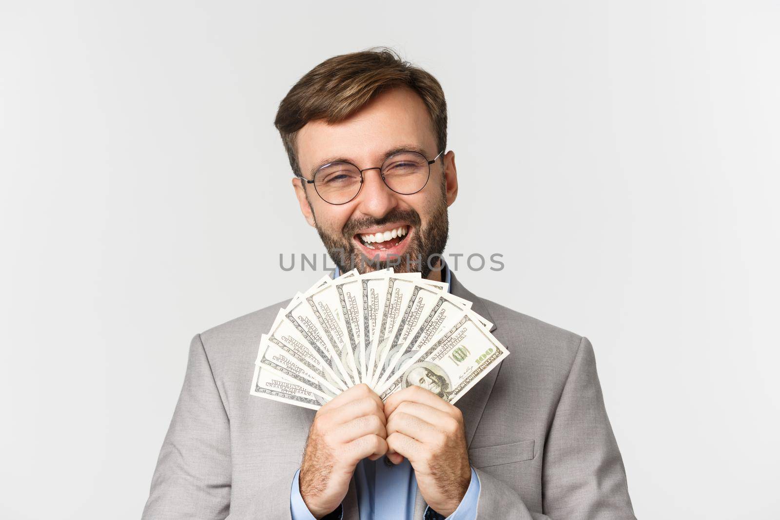 Close-up of rich and successful businessman, wearing gray suit and glasses, showing cash and laughing accomplished, standing over white background.