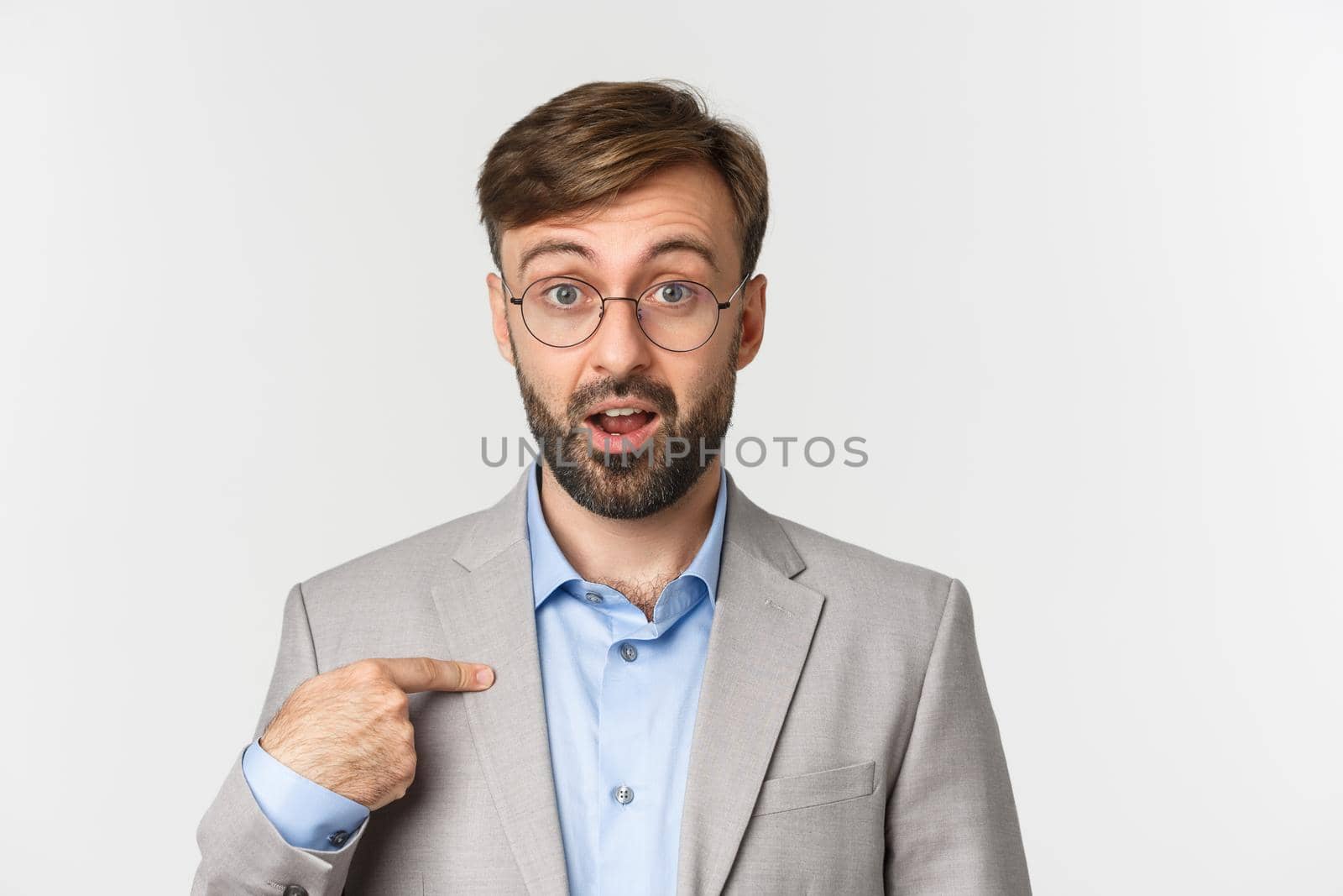 Close-up of surprised and excited office worker in glasses and gray suit, pointing finger at camera and looking confused, standing over white background.