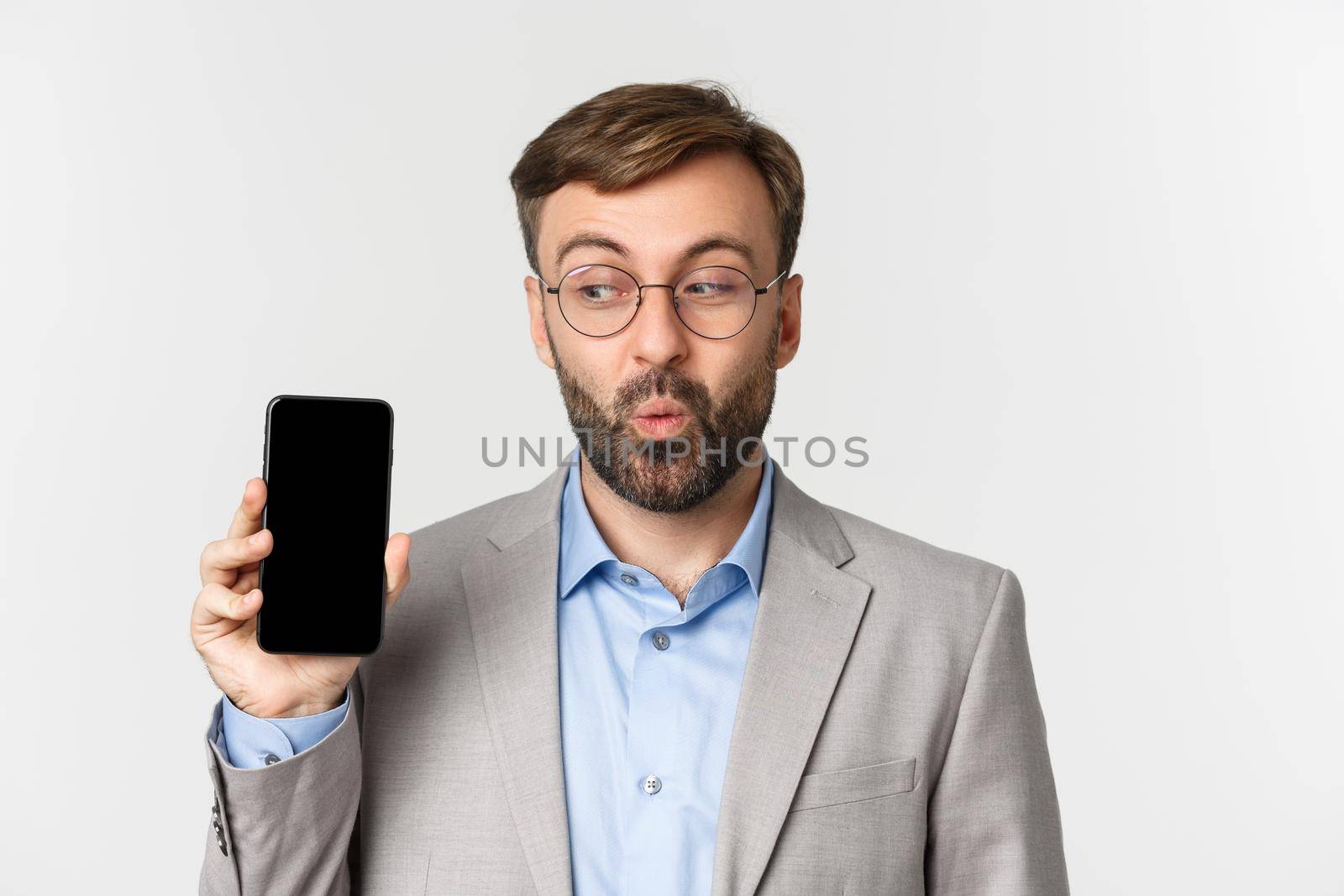 Close-up of excited businessman in gray suit and glasses, showing mobile phone screen and looking happy, standing over white background.