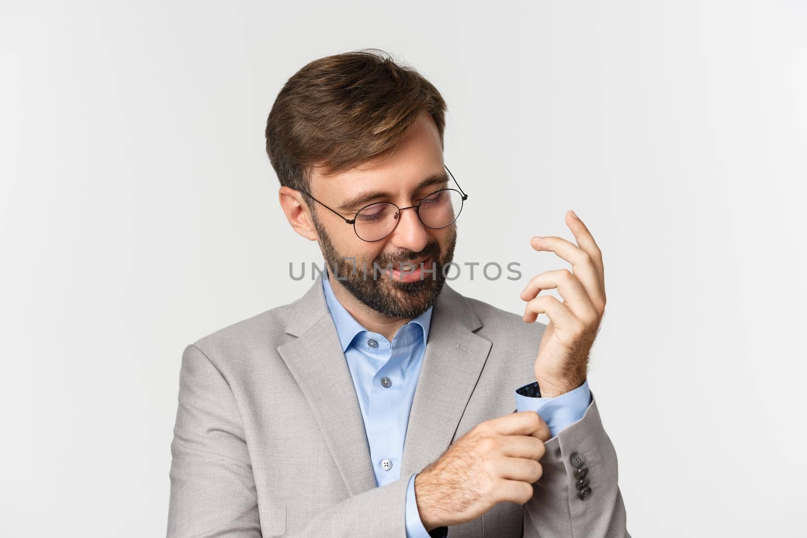 Close-up of confident businessman in glasses getting dressed for office, buttoning sleeve and smiling, standing over white background.