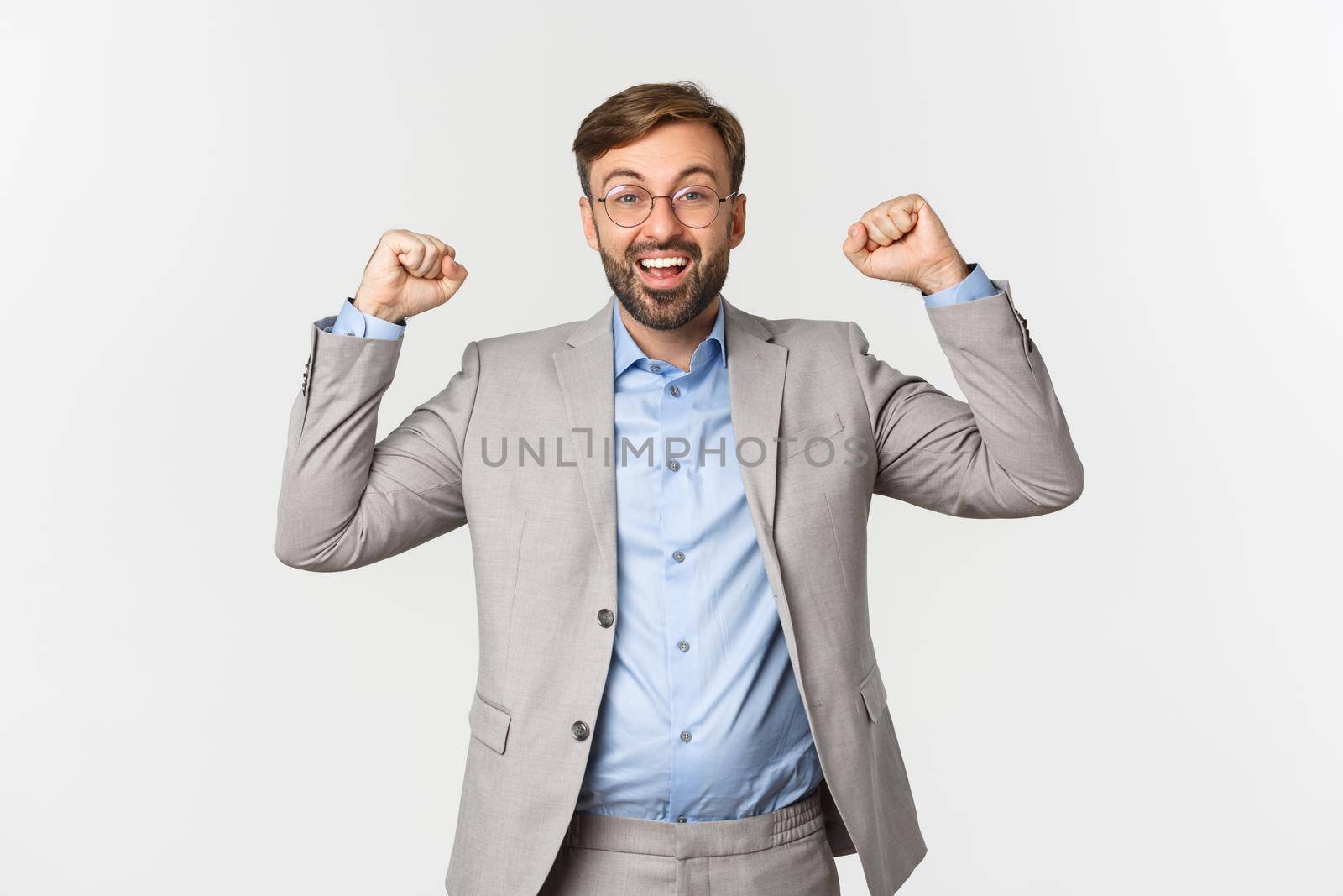 Portrait of happy businessman with beard, wearing grey suit and glasses, raising hands up in hooray gesture, saying yes and rejoicing, celebrating victory or achievement, white background.