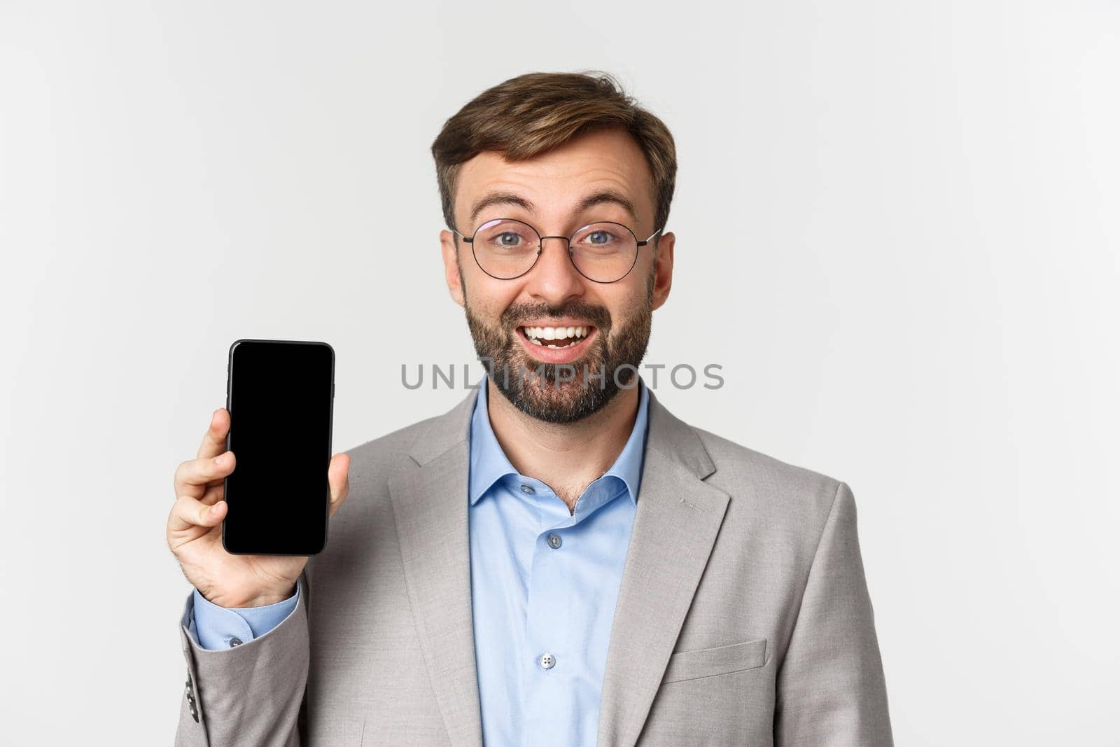 Close-up of cheerful male entrepreneur in glasses and gray suit, showing smartphone screen and smiling, standing over white background.