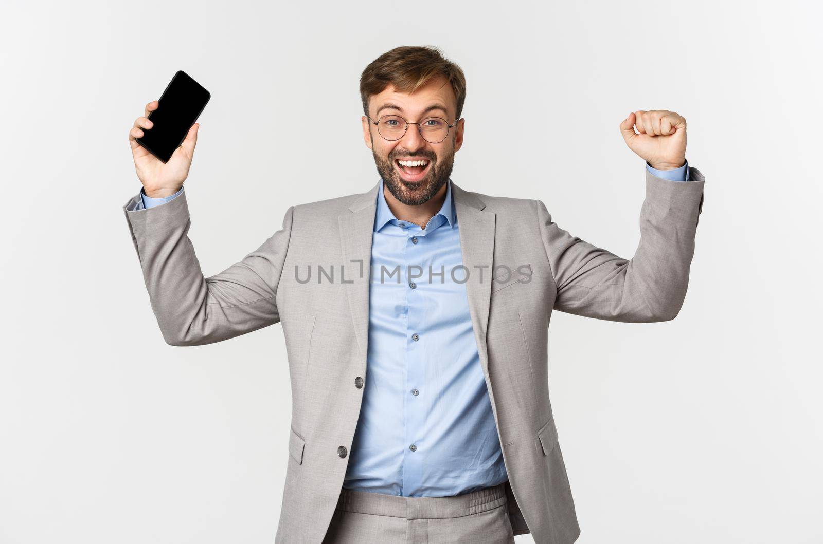 Image of successful businessman in grey suit and glasses, holding mobile phone, raising hands up and shouting yes from satisfaction, triumphing over white background by Benzoix