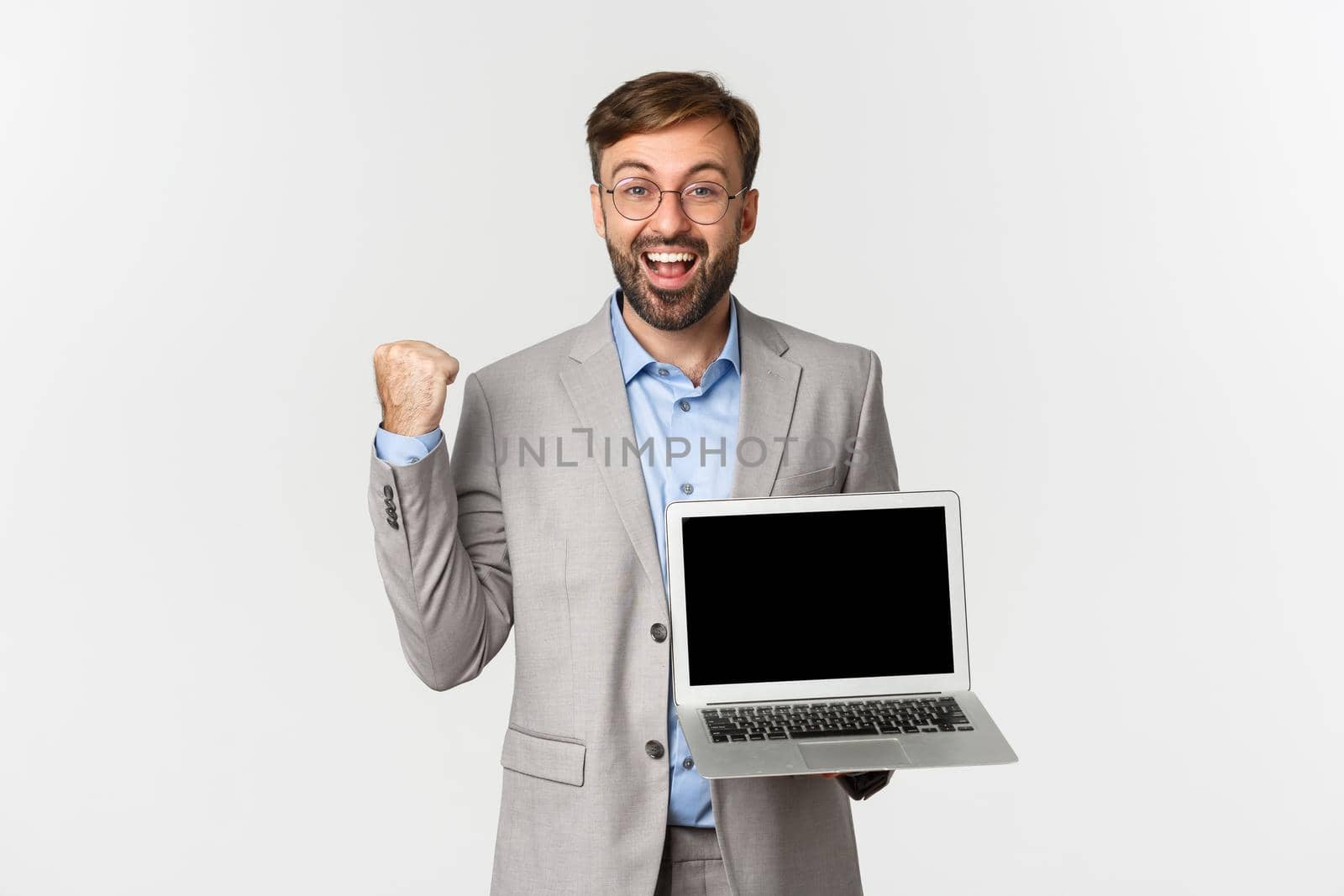 Portrait of happy bearded businessman in grey suit and glasses, showing his accomplishment on laptop screen, rejoicing and saying yes, demonstrating presentation, white background.