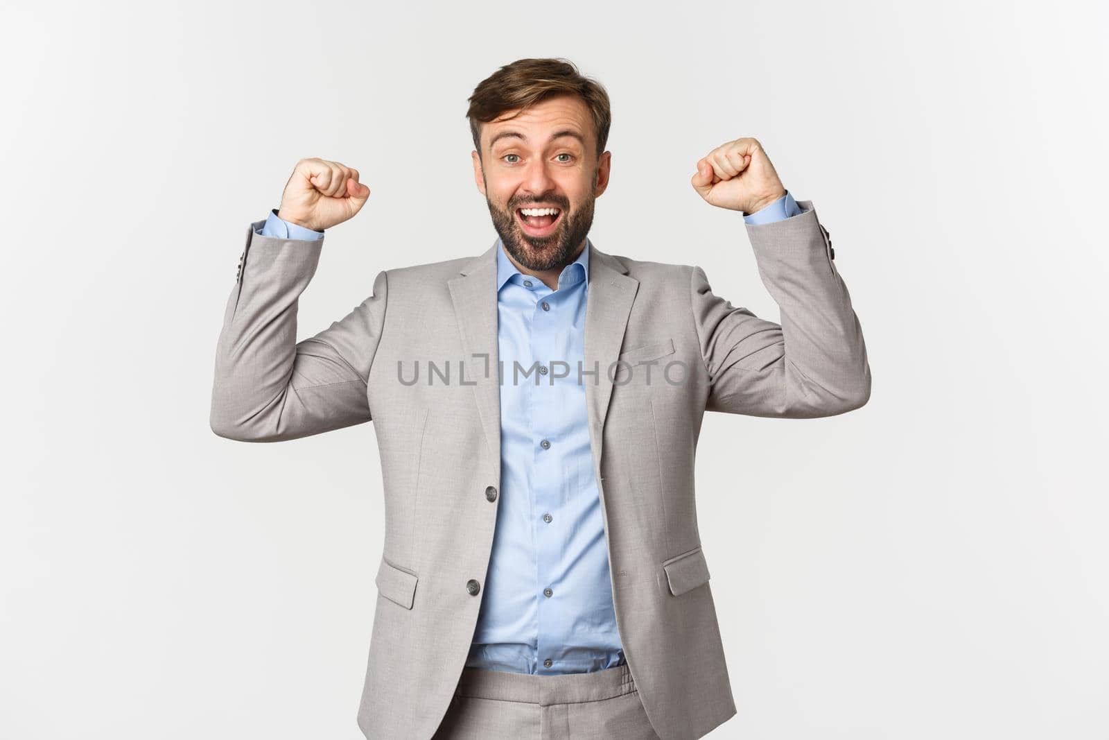 Portrait of relieved and happy bearded man in business suit, rejoicing over win, raising hands up and saying yes with satisfaction, standing over white background by Benzoix