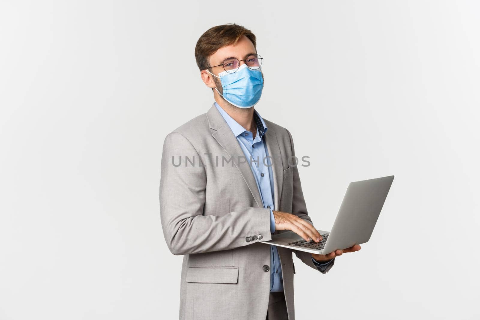 Concept of work, covid-19 and social distancing. Image of working businessman in medical mask and suit, using laptop and smiling happy, standing over white background by Benzoix