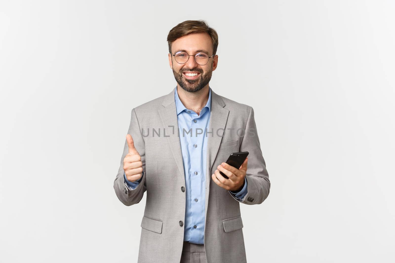 Portrait of handsome successful businessman with beard, wearing glasses and gray suit, holding mobile phone and showing thumbs-up in approval, smiling pleased, white background by Benzoix