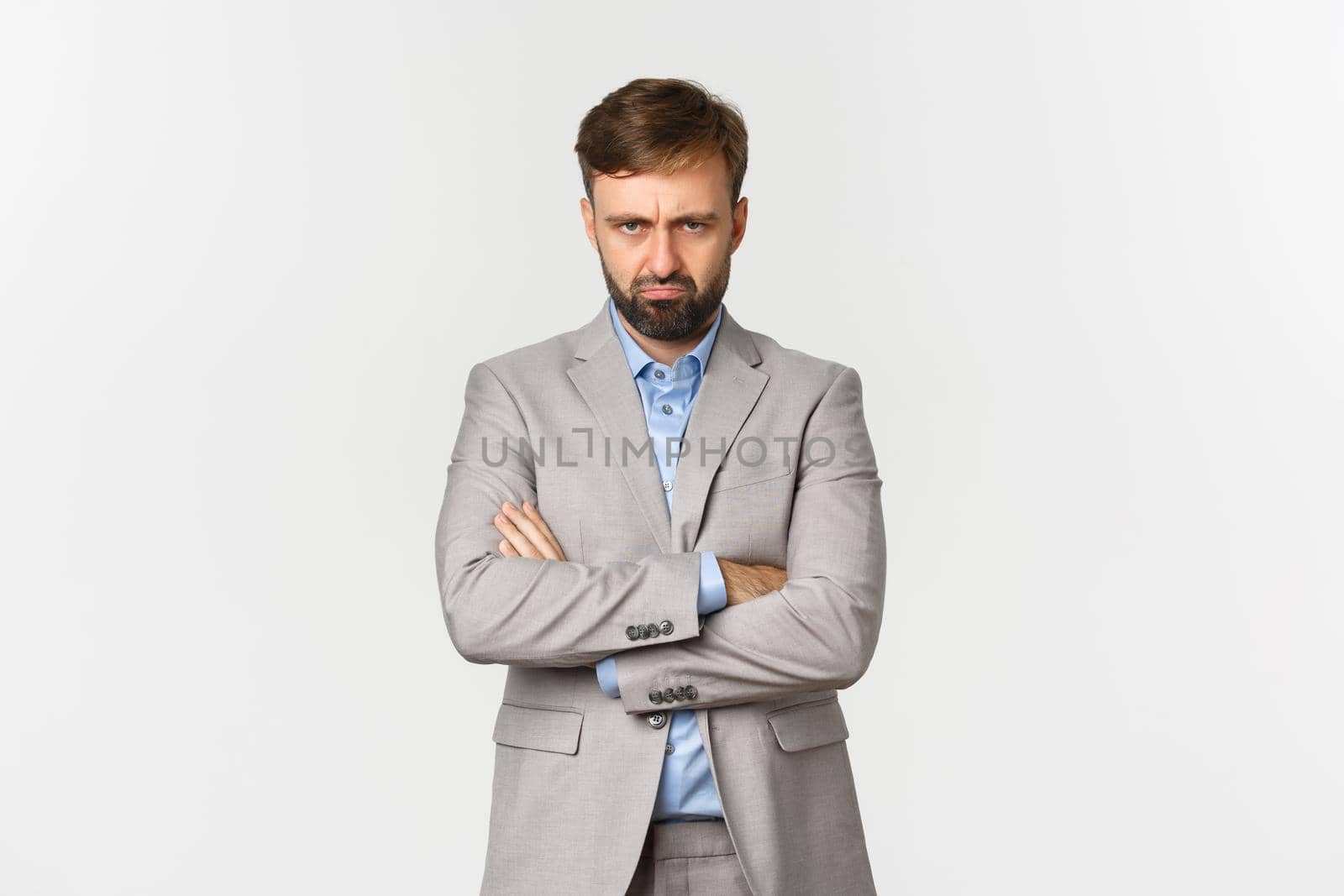 Portrait of offended gloomy bearded guy in grey suit, sulking and standing with arms crossed over chest, angry at someone, posing over white background.