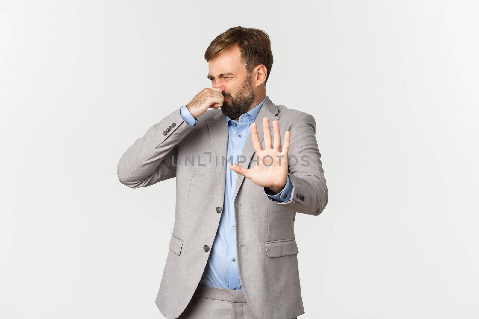 Portrait of disgusted bearded businessman in grey suit, rejecting something with awful smell, shut nose and making decline sign, standing over white background.