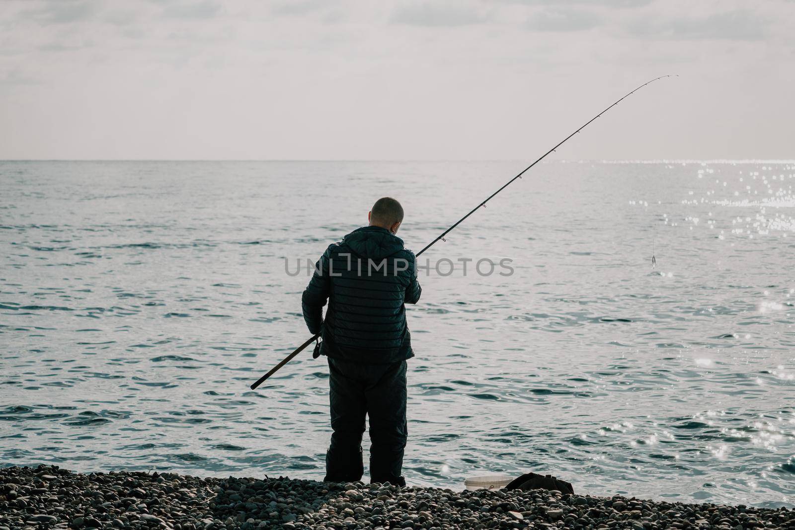 Man hobby fishing on sea tightens a fishing line reel of fish. Calm surface sea. Close-up of a fisherman hands twist reel with fishing line on a rod. Fishing in the blue sea outdoors. by panophotograph