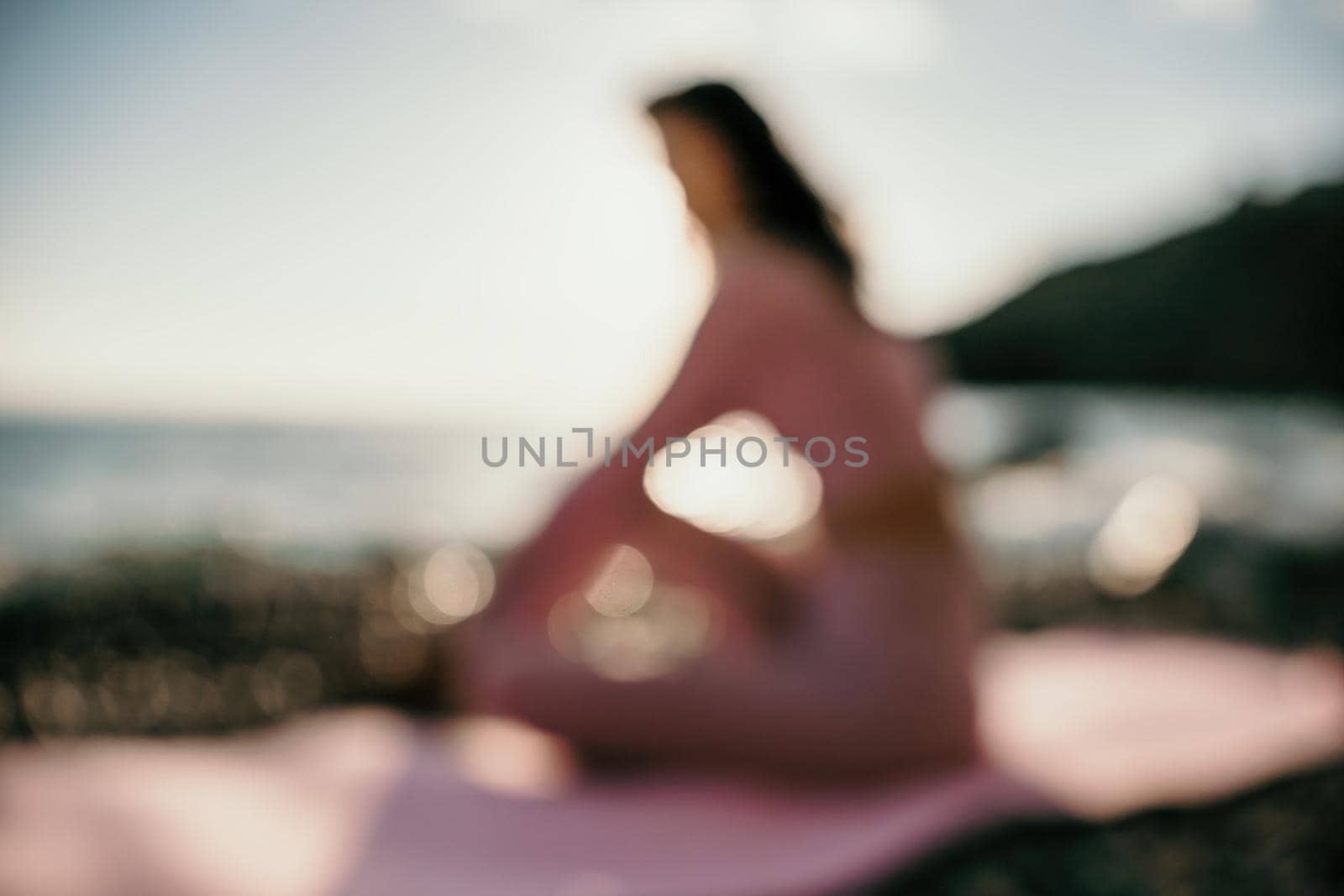 Abstract defocused silhoute of middle aged well looking woman with black hair doing Pilates with the ring on the yoga mat near the sea on the pebble beach. Female fitness yoga concept. by panophotograph