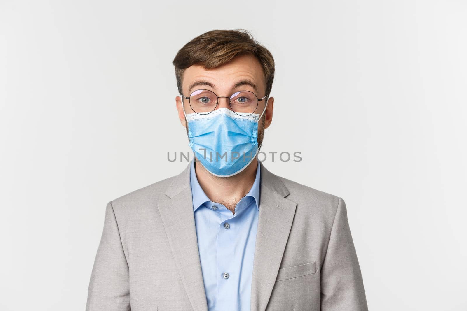 Concept of work, covid-19 and social distancing. Close-up of surprised man in gray suit and medical mask, raising eyebrows and looking amazed at camera, standing over white background by Benzoix