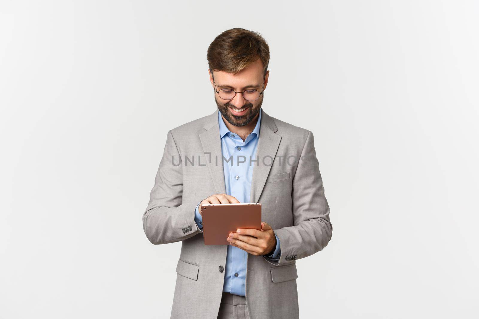 Image of businessman working, looking at digital tablet and smiling pleased, standing in grey suit and glasses over white background by Benzoix