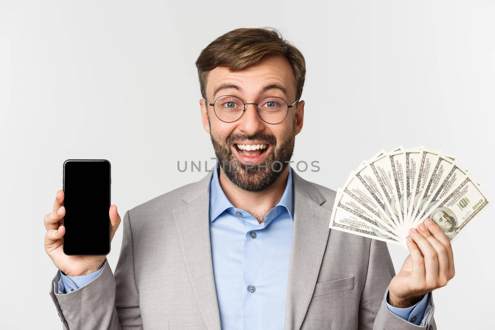 Close-up of successful businessman in gray suit and glasses, showing money and smartphone screen, smiling cheerful, standing over white background.
