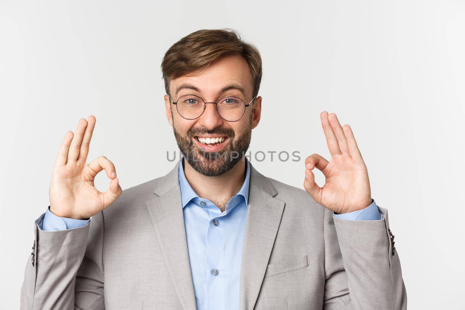 Close-up of handsome bearded man in gray suit and glasses, recommend something, showing okay sign and smiling, approve good choice, standing over white background by Benzoix